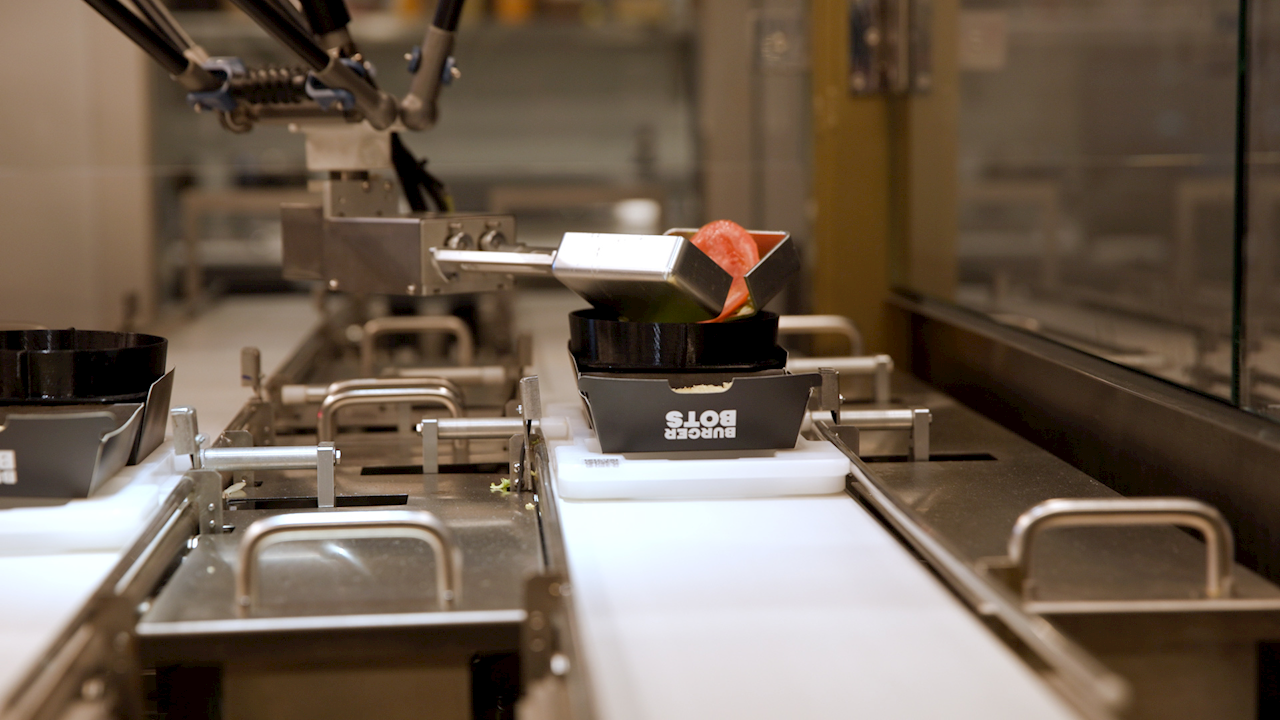 Braccio robotico BurgerBots che aggiunge ingredienti su un burger in linea